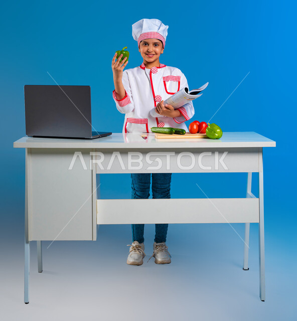 أماني وأحلام الطفولة في الحصول على مهنة الشيف, بورتريه مقرب لفتاة عربية خليجية سعودية ترتدي زي الطباخة تمسك كتاب الطهي وتنظر إلى الكاميرا, فوائد استخدام الفلفل الأخضر في الطبخ, إعداد وتحضير الأكلات الصحية, الشغف في تعلم وصفات جديدة عبر الانترنت, خلفية زرقاء