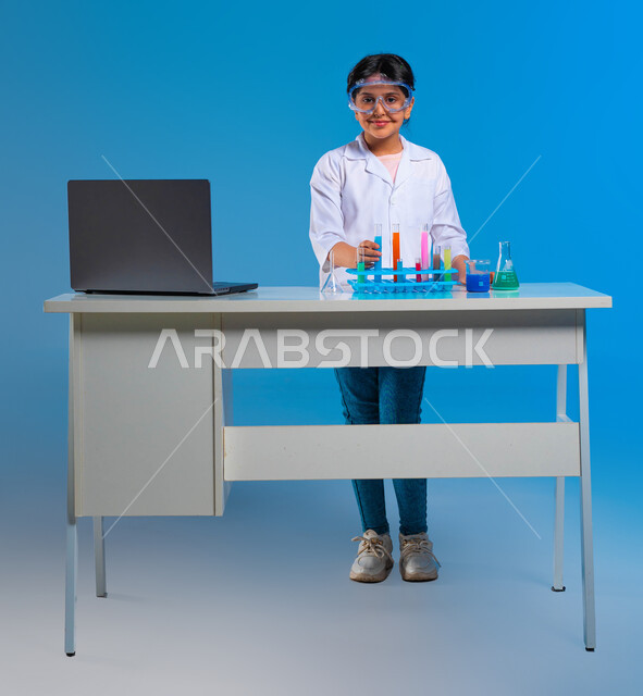 استخدام أنابيب الاختبار والقيام بالتفاعلات والأبحاث العلمية, الوقوف بإيماءات الثقة والبشاشة, شغف التعلم وحلم الحصول على مهنة الباحثة الكيميائية, بورتريه لفتاة عربية خليجية سعودية ترتدي المعطف الأبيض ونظارة الحماية تنظر إلى الكاميرا, خلفية زرقاء
