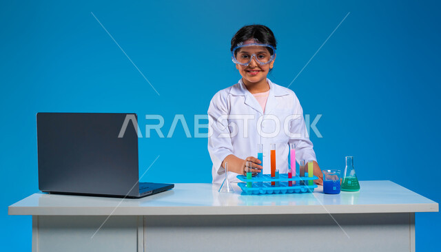 تدوين الملاحظات عبر جهاز الحاسوب, شغف التعلم وحلم الحصول على مهنة الباحثة الكيميائية, بورتريه مقرب لفتاة عربية خليجية سعودية ترتدي المعطف الأبيض ونظارة الحماية تنظر إلى الكاميرا, استخدام أنابيب الاختبار والقيام بالتفاعلات والأبحاث العلمية, خلفية زرقاء
