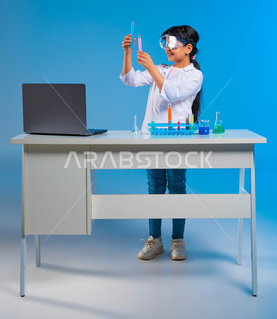 تحقيق حلم الطفولة في الوصول إلى مهنة الباحثة الكيميائية, إجراء تجارب علمية في معمل الكيمياء, بورتريه لفتاة عربية خليجية سعودية ترتدي المعطف الأبيض ونظارة الحماية تستخدم أنابيب الاختبار, القيام بالبحوث والتفاعلات في المختبرات التعليمية, خلفية زرقاء