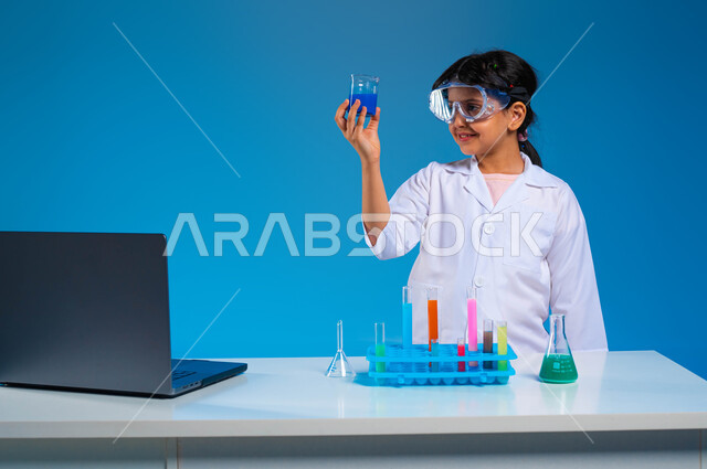 اجراء تجارب وتفاعلات كيميائيه حيويه في المختبر، بورتريه مقرب لفتاة عربية خليجية سعودية ترتدي معطف أبيض وتستخدم انابيب الاختبار، أبحاث علمية في معمل الكيمياء، أمنية الحصول على مهنة الباحثة الكيميائية، استخدام جهاز الحاسوب لتدوين الملاحظات والنتائج الطبيه، خلفية زرقاء
