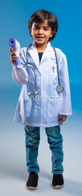 اتباع إجراءات الوقاية والسلامة، بورتريه  لولد عربي خليجي سعودي يرتدي المعطف الطبي الأبيض وسماعة الطبيب ويحمل بيده ميزان الحرارة الرقمي، مفهوم الطب والرعاية الصحية، اختيار وتحديد المهنة المستقبلية منذ الصغر، صورة الجسم الكامل، خلفية زرقاء