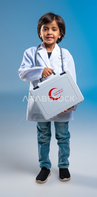 Dealing with emergency situations, the concept of health care and medicine, a portrait of a smiling Saudi Arabian Gulf boy wearing a white medical coat and stethoscopes and holding a first aid bag in his hand, future goals and wishes to reach the profession of emergency medicine, full-length body portrait, blue background