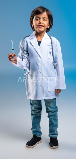 تحديد واختيار مهنة المستقبل منذ الصغر، مفهوم الطب والرعاية الصحية، بورتريه  لولد عربي خليجي سعودي مبتسم يرتدي المعطف الطبي الأبيض وسماعات الطبيب ويحمل بيده حقنة طبية، صورة الجسم بالطول الكامل، أهداف مستقبيله في العمل في المختبرات والتحاليل الطبيه، خلفية زرقاء