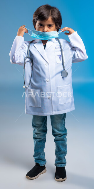 صورة الجسم بالطول الكامل، نظرات الجد والاجتهاد للوصول إلى مهنة الطب، مفهوم الطب والرعاية الصحية، بورتريه  لولد عربي خليجي سعودي يرتدي المعطف الطبي الأبيض وسماعات الطبيب ويقوم بوضع الكمامة، اختيار وتحديد المهنة المستقبلية منذ الصغر، خلفية زرقاء