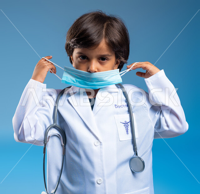 طموحات واهداف مستقبلية، مفهوم الطب والرعاية الصحية، بورتريه مقرب لولد عربي خليجي سعودي يرتدي المعطف الطبي الأبيض وسماعات الطبيب ويقوم بوضع الكمامة، نظرات الجد والاجتهاد للوصول الى مهنة الطب، خلفية زرقاء
