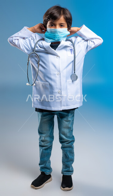 Choosing and defining a future profession since childhood, future ambitions and goals, the concept of medicine and health care, a portrait of a Saudi Arabian Gulf boy wearing a white medical coat and wearing a mask and stethoscopes, full-length body portrait, blue background.