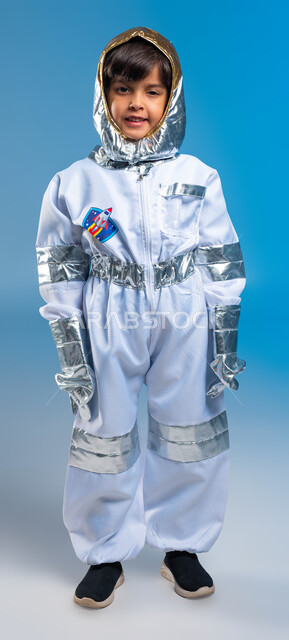 تطور و دعم قطاع الفضاء في المملكة العربية السعودية، بورتريه لولد عربي سعودي خليجي مبتسم يرتدي زي رائد الفضاء، نظرات الشغف و الجد لتحقيق أهداف في المستقبل، حلم الوصول إلى القمر، رواد فضاء سعوديين، صورة للجسم كامل، خلفية زرقاء .