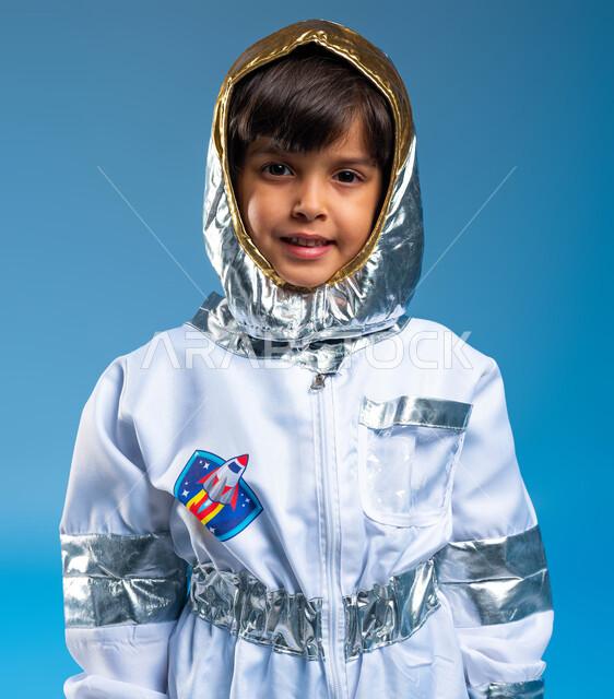He dreams of becoming an astronaut, a close-up portrait of a smiling Saudi Gulf Arab boy wearing an astronaut uniform, gestures of seriousness and effort to achieve a childhood dream, wishes and hopes for future Saudi careers, the development of space science and astronomy in the Kingdom of Saudi Arabia, a blue background.