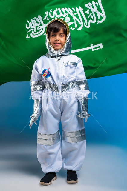 ذكرى اليوم الوطني ٢٣ سبتمبر، بورتريه لولد عربي سعودي خليجي مبتسم يرتدي زي رائد الفضاء، يوم العلم السعودي ١١ مارس، نظرات الجد و الاجتهاد للوصول إلى الفضاء و القمر، مهن و وظائف سعودية مستقبلية، صورة للجسم كامل، خلفية زرقاء .