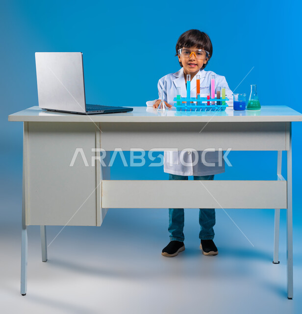 امنيات مستقبلية كطبيب مختبرات كيميائيه، بورتريه لولد عربي سعودي خليجي مبتسم يرتدي معطف أبيض و يضع نظارات واقية لإجراء التفاعلات الحيوية، أبحاث علمية باستخدام أنابيب الأختبار، تدوين النتائج و الملاحظات المخبرية على الحاسوب، صورة للجسم كامل، خلفية زرقاء .