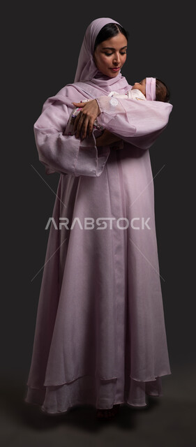 التعامل مع التحديات والضغوط عند تربية الطفل الرضيع، مفهوم الطفولة والامومة، تهدئة الأطفال الصغار وتخفيف التوتر والقلق لديهم من خلال الاحتضان، بورتريه لامرأة عربية خليجية سعودية محجبة ترتدي العباءة وتحمل ابنتها بحب وحنان، صورة الجسم بالطول الكامل، خلفية سوداء