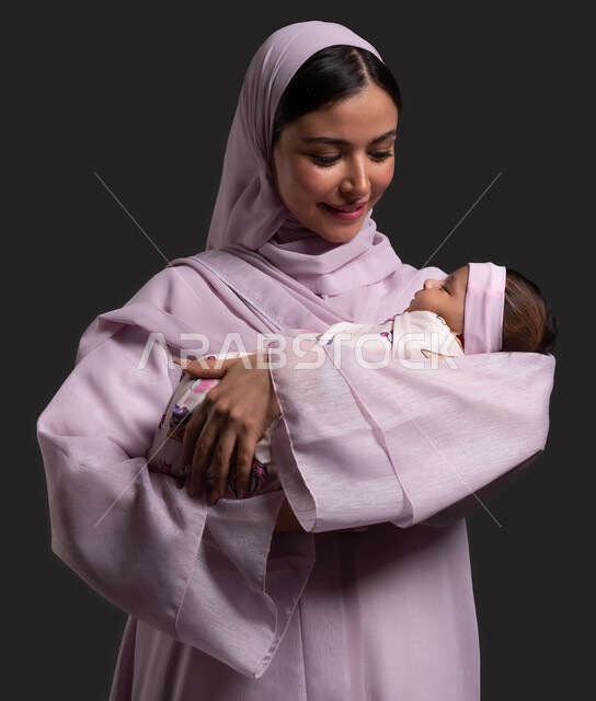 تقديم الأمان والاستقرار للطفل، استمداد الرضيع الطمأنينة والاستقرار من الأم، بورتريه مقرب لإمرأة عربية خليجية سعودية محجبة ترتدي العباءة وتحتضن طفلتها، الاستمتاع بقضاء وقت ممتع مع الأطفال الرضع، حنان الأمهات والعناية بالأبناء، خلفية سوداء