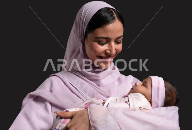 الاستمتاع باللحظات الجميلة مع الطفل ، بناء علاقة قوية ومتينة مع الابناء، التضحية والتفاني من أجل سعادة وراحة الرضيع،  بورتريه مقرب لأم عربية خليجية سعودية محجبة ترتدي العباءة تضم ابنتها الى صدرها وتضحك بسعادة، خلفية سوداء