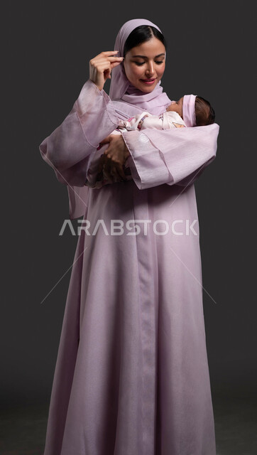 التعبير عن الحب والاهتمام، لفت الانتباه بحركات اليدين، مداعبة الطفلة بلطف وحنان، بورتريه لإمرأة عربية خليجية سعودية محجبة ترتدي العباءة وتلاعب طفلتها بحب وحنان، بناء علاقة قوية ومتينة مع الطفل الرضيع، خلفية سوداء