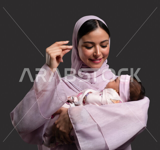 تحمل الام مسؤولية رعاية وتربية الرضيعة، لفت الانتباه بحركات اليدين، مداعبة الطفلة بلطف وحنان،  حنان الأمهات والعناية بالأبناء، دور الأم تجاه أولادها، بورتريه مقرب لإمرأة عربية خليجية سعودية محجبة ترتدي العباءة وتقضي وقت ممتع مع طفلتها، خلفية سوداء