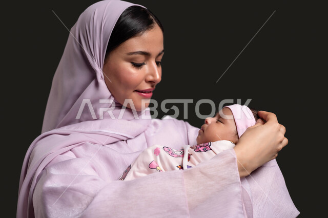 الدور الأساسي للأم في رعاية أبناءها، مشاعر الأمومة والطفولة، تحمل المسؤولية والعناية بالأطفال، بورتريه لإمرأة عربية خليجية سعودية ترتدي العباءة الوردية تحمل صغيرتها وتنظر إليها بحب وحنان، خلفية سوداء