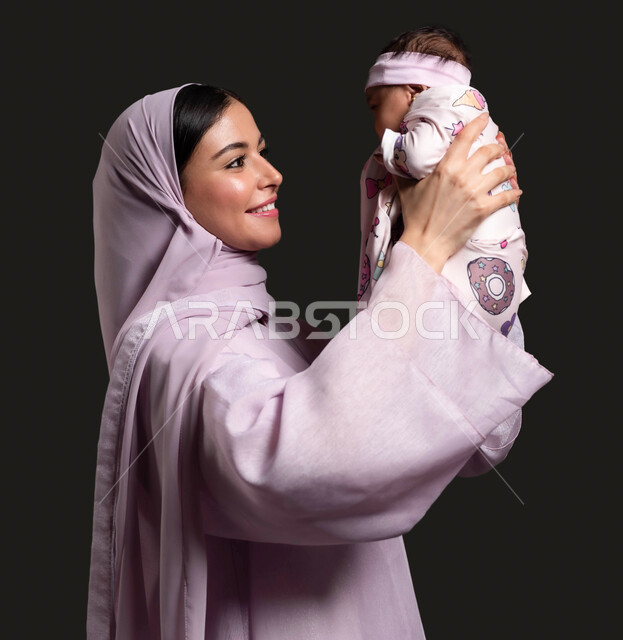 رعاية وتربية الطفل الرضيع، حنان الأمهات والعناية بالأطفال، دور الأم تجاه أولادها، الاستمتاع واللعب مع الأبناء، بورتريه مقرب لسيدة عربية خليجية سعودية محجبة ترتدي العباءة وتقضي وقت ممتع مع طفلتها، خلفية سوداء