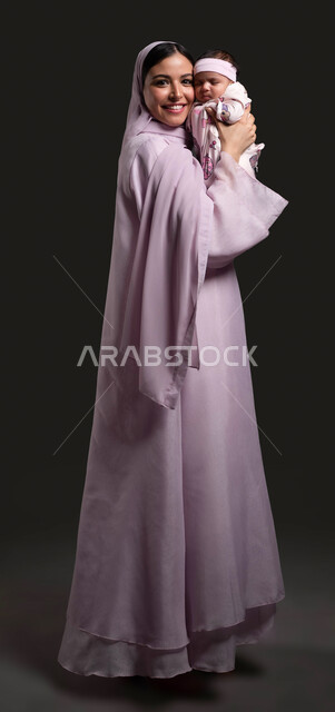 بناء علاقة قوية ومتينة مع الرضيعة، نظر الى الكاميرا، التضحية والتفاني من أجل سعادة وراحة الطفل، نظر الى الكاميرا، حنان الأمهات والعناية بالأطفال، دور الأم تجاه أولادها، بورتريه مقرب لسيدة عربية خليجية سعودية محجبة ترتدي العباءة وتضم ابنتها الى صدرها، صورة الجسم بالكامل، خلفية سوداء