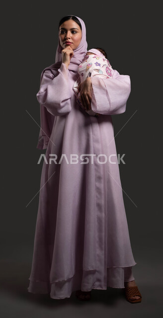 التخطيط  لمستقبل الطفلة الرضيعة، التمعن والتأمل في موضوع ما، ايماءات التفكير والتركيز، بورتريه مقرب لسيدة عربية خليجية سعودية محجبة ترتدي العباءة الملونة وتضع يدها على ذقنها بايماءات الحيرة، صورة لكامل الجسم، خلفية سوداء
