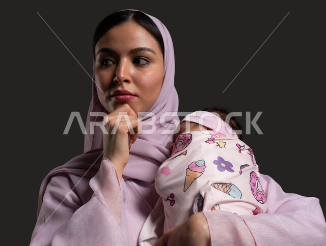 Gestures of contemplation and mind wandering, contemplating a topic, planning for the baby girl’s future, a close-up portrait of a veiled Saudi Arabian Gulf mother wearing a colorful abaya and placing her hand on her chin with contemplative gestures, black background.