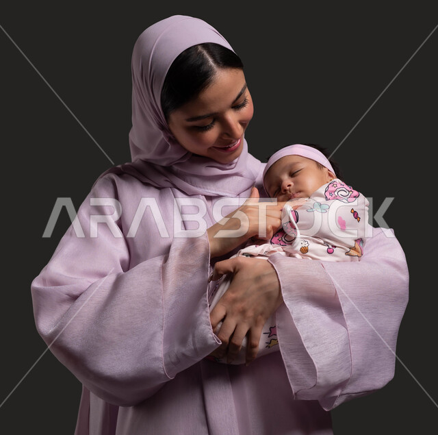 تهدئة الأطفال الصغار وتخفيف التوتر والقلق لديهم من خلال الاحتضان، بورتريه مقرب لسيدة عربية خليجية سعودية محجبة ترتدي العباءة الملونة وتنظر الى رضيعتها بحب وحنان، تقوية علاقة الثقة والود بين الأم والطفل، خلفية سوداء