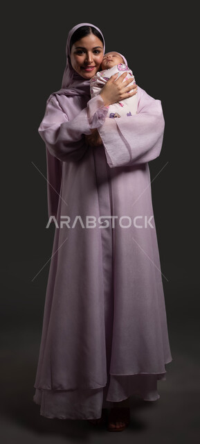 تقديم الدعم العاطفي للأطفال، بورتريه لسيدة عربية خليجية سعودية محجبة ترتدي العباءة الملونة تحتضن طفلتها النائمة وتنظر الى الكاميرا بسعادة، تلبية احتياجات الأبناء والعناية بهم، صورة الجسم بالكامل، خلفية سوداء