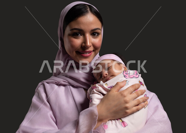 تربية الأولاد ورعايتهم، تعزيز رابط الأمومة بين الأم وصغيرتها، مفهوم حنان الأمهات و تحمل المسؤولية والعناية بالأطفال، بورتريه مقرب لأم عربية خليجية سعودية تحمل طفلتها الرضيعة وتنظر إلى الكاميرا، الشعور بالأمان والاطمئنان، خلفية سوداء.
