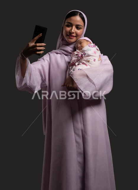 تحسين جودة الحياة باستخدام التقنية الحديثة ، توثيق اللحظات السعيدة مع الرضيعة، بورتريه لأم عربية خليجية سعودية مبتسمة ترتدي العباءة تضم صغيرتها المقمطة وتلتقط معها صورة سيلفي عبر الهاتف المحمول، مشاعر الحب والحنان بين الأم وأولادها، خلفية سوداء.