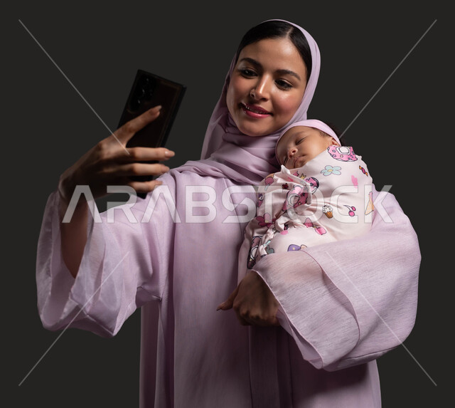 التقاط صور السليفي التذكارية عبر الهاتف المحمول، توثيق اللحظات السعيدة مع الرضيعة، بورتريه لأم عربية خليجية سعودية مبتسمة ترتدي العباءة تحتضن طفلتها المقمطة بحب وحنان، براءة الأطفال وبساطتهم، استخدام الأجهزة والتقنيات الحديثة، خلفية سوداء.