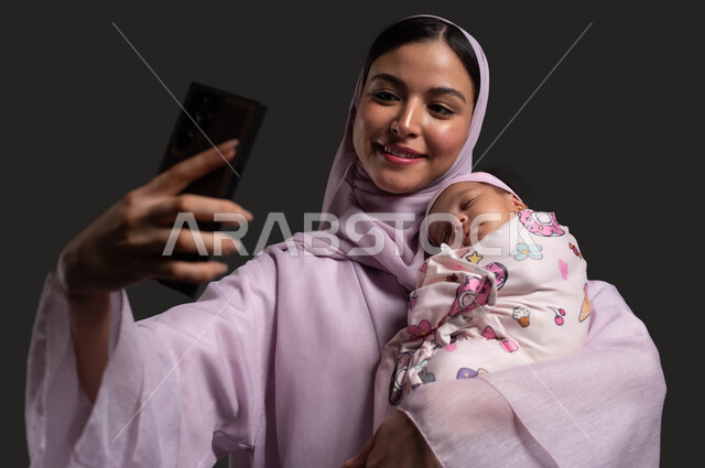 توثيق اللحظات السعيدة مع الرضيعة، التقاط صور السليفي التذكارية عبر الهاتف المحمول، بورتريه مقرب لأم عربية خليجية سعودية مبتسمة ترتدي العباءة تحمل صغيرتها المقمطة بحب وحنان، التعامل مع الأطفال الرضع، استخدام الأجهزة والتقنيات الحديثة، خلفية سوداء.