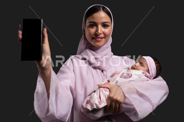 استعمال التقنيات الحديثة في التسويق الالكتروني، بورتريه مقرب لأم عربية خليجية سعودية محجية ترتدي العباءة الملونة تحمل ابنتها المقمطة في يدها وتمسك الجوال بشاشة سوداء فارغه، استخدام الهاتف المحمول للبحث عن معلومات الامومه والرعايه الصحيه للأطفال، خلفية سوداء
