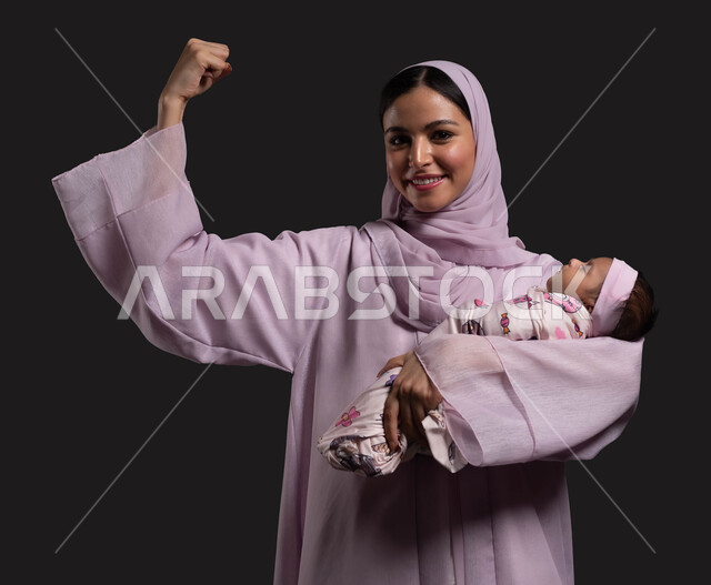 ايماءات الراحة والانتصار، تربية الأطفال والعناية بهم، دور ومسؤوليه الأم تجاه أولادها، بورتريه مقرب لسيدة عربية خليجية سعودية محجبة ترتدي العباءة الملونة تحتضن طفلتها النائمة وترفع قبضة يدها للأعلى، خلفية سوداء