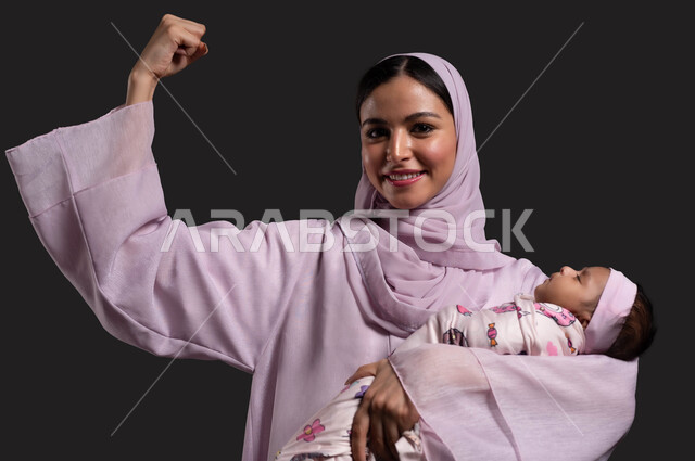تقوية رابطة الأمومة بين الأم وطفلتها، بورتريه مقرب لأم عربية خليجية سعودية محجبة ترتدي العباءة الملونة تحمل طفلتها النائمة، ايماءات السعادة بالنجاح و الانجاز، الحاجة للراحة والنوم، تحقيق الهدف في تهدئة ونوم الطفلة، خلفية سوداء