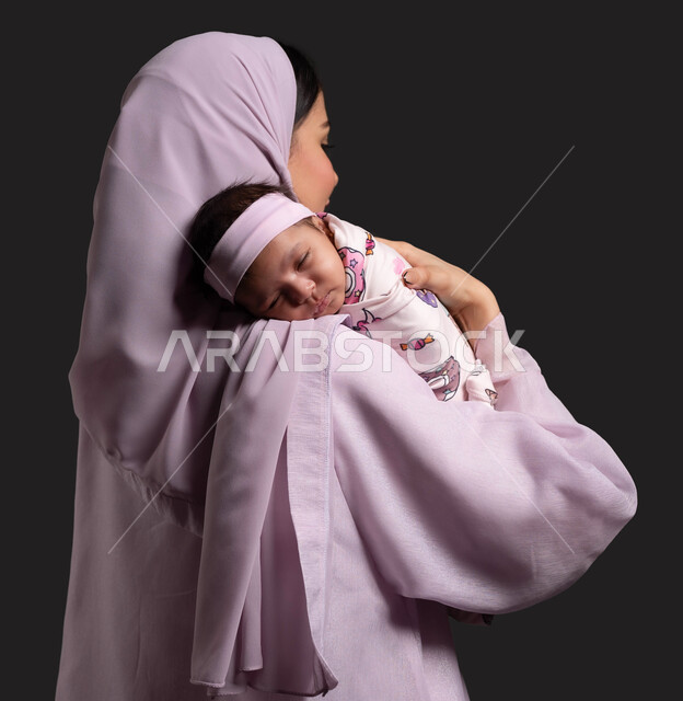 دور الأم في حياه الأبناء، بورتريه مقرب من الخلف لأم عربية خليجية سعودية محجبة ترتدي العباءة وتحتضن ابنتها بين ذراعيها، مفهوم الحب والحنان، مساعدة الرضيعة على الاسترخاء والنوم، تقوية رابطة الأمومة بين الأم وطفلتها، خلفية سوداء