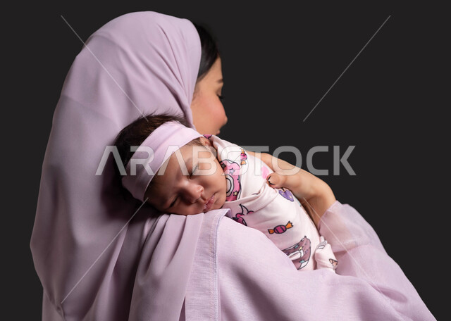 التعامل مع الأطفال الرضع، بورتريه مقرب من الخلف لأم عربية خليجية سعودية محجبة ترتدي العباءة الملونة و تحمل رضيعتها النائمة بحب وحنان، مساعدة الرضيعة على الاسترخاء والنوم، مفهوم الامومة والطفولة، خلفية سوداء