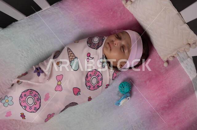 تقميط الأطفال بالبطانيات، إيماءات السعادة والأمان،صورة علوية عمودية لطفلة رضيعة عربية خليجية سعودية نائمة ملفوفة بغطاء مورد باللون الزهري، أهمية الراحة والنوم، الشعور بالتعب والنعاس، مفهوم البراءة والطفولة