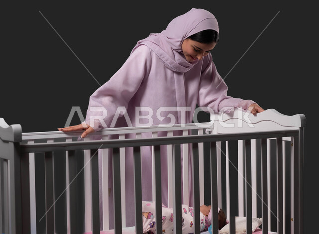 قضاء أوقات جميلة مع الاطفال، بورتريه لأم عربية خليجية سعودية محجبة ترتدي العباءة تقف بجانب سرير طفلتها الرضيعة للاطمئنان عليها، مشاعر الحب والحنان بين الأم وأولادها، الاستمتاع بمشاعر الأمومة، خلفية سوداء