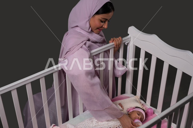حنان الأم ورعايتها لأطفالها، بورتريه مقرب لام عربية خليجية سعودية محجبة ترتدي عباءة ملونة تقوم بإطعام طفلتها الرضيعة بإستخدام زجاجة الرضاعة، إيماءات الراحة والأمان، مفهوم الرضاعة الصناعية، خلفية سوداء