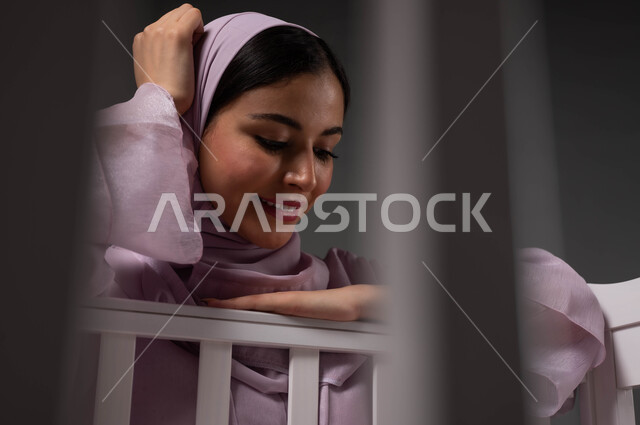 إيماءات الرضا بالسهر على راحة الرضيعة، تقوية رابط الأمومة والبنوة، الاستمتاع بقضاء الوقت مع الأبناء، الشعور بالأمان والاطمئنان، صورة مقربة لإمرأة عربية خليجية سعودية محجبة مبتسمة تنظر إلى صغيرتها بإيماءات الحب، أجواء الراحة والهدوء