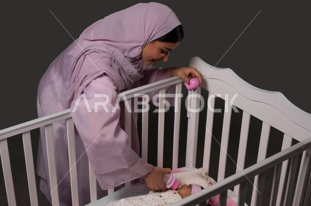 الاستلقاء في المهد وتناول الوجبات الصحية، استخدام عبوة الرضاعة لشرب الحليب، حنان الأم ورعايتها لأطفالها، بورتريه لسيدة عربية خليجية سعودية محجبة ترتدي العباءة تعطي رضيعتها الغذاء المفيد، إيماءات الراحة والأمان، مفهوم الرضاعة الصناعية، خلفية سوداء