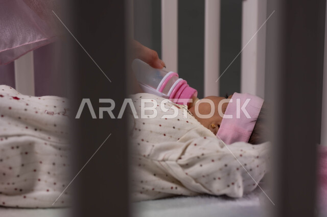 مفهوم الرضاعة الصناعية، استخدام عبوة الرضاعة لشرب الحليب، حنان الأم ورعايتها لأبناءها، صورة مقربة لطفلة عربية خليجية سعودية مستلقية في المهد تتناول الغذاء المفيد، إيماءات الراحة والأمان .