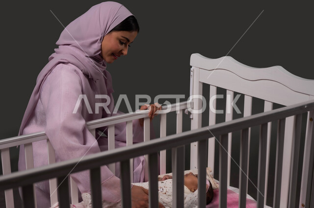 Caressing infants gently and calmly, feeling safe and reassured, enjoying spending time with children, strengthening the bond of motherhood and sonship, portrait of a veiled Saudi Gulf Arab woman sitting next to the bed and looking at her little girl with gestures of love, black background