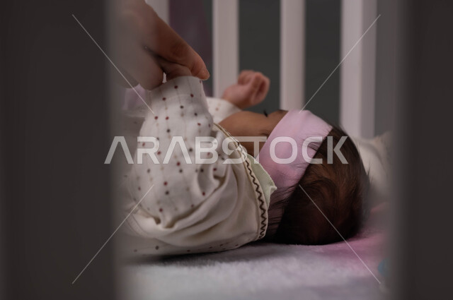المحافظة على هدوء الطفلة، الاسترخاء والشعور بالأمان، مداعبة الرضع بلطف وحنان، الاستمتاع بقضاء الوقت مع الأبناء، صورة مقربة لرضيعة عربية خليجية سعودية تستلقي في المهد تمسك والدتها يدها، ملامح ناعمة وبريئة .
