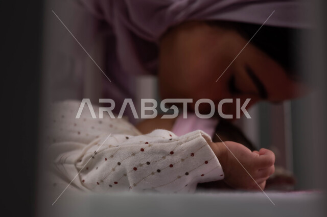 تقوية رابط الأمومة والبنوة، ملامح ناعمة وبريئة, الاسترخاء والنوم بعمق، الشعور بالأمان والاطمئنان، صورة مقربة لإمرأة عربية خليجية سعودية محجبة تغمض عينيها تقبل رضيعتها في السرير بإيماءات الحب، أجواء الراحة والهدوء .