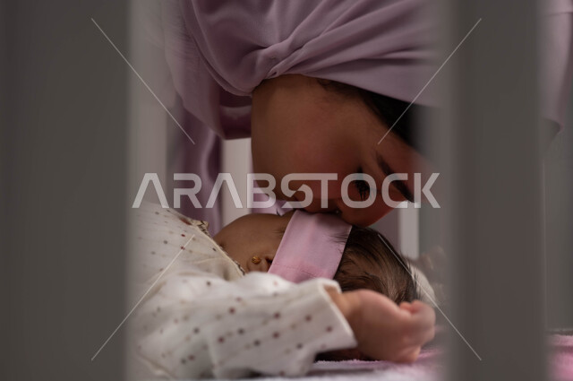 الاسترخاء والنوم بعمق، الشعور بالأمان والاطمئنان، تقوية رابط الأمومة والبنوة، صورة مقربة لإمرأة عربية خليجية سعودية محجبة تغمض عينيها تقبل رضيعتها في السرير بإيماءات الحب، أجواء الراحة والهدوء، ملامح ناعمة وبريئة .
