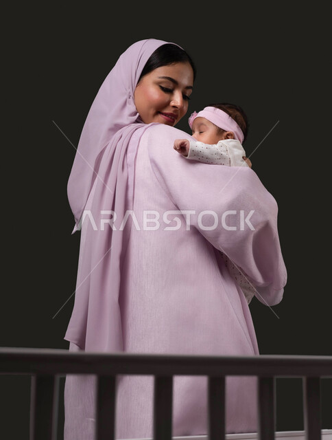 Dealing with infants gently, standing in front of the bed and looking at the infant, the concept of motherhood and childhood, playing with children with affection and care, a close-up portrait of a smiling Saudi Arabian Gulf woman wearing an abaya carrying her little girl and caressing her with love and tenderness, black background