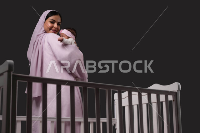 التعامل مع الأطفال الرضع برفق، الوقوف أمام السرير والنظر إلى الكاميرا، مفهوم الأمومة والطفولة، ملاعبة الأبناء بعطف واهتمام، بورتريه لأم عربية خليجية سعودية تبتسم ترتدي العباءة تحمل صغيرتها وتقوم بمداعبتها بحب وحنان, خلفية سوداء