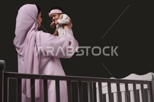 الوقوف أمام السرير واللعب مع الأبناء، حنان الأمهات و تحمل المسؤولية والعناية بالأطفال، ملاعبة البنات بعطف واهتمام، بورتريه لأم عربية خليجية سعودية تبتسم ترتدي العباءة تحمل طفلتها الرضيعة وتقوم بمداعبتها بحب وحنان, خلفية سوداء