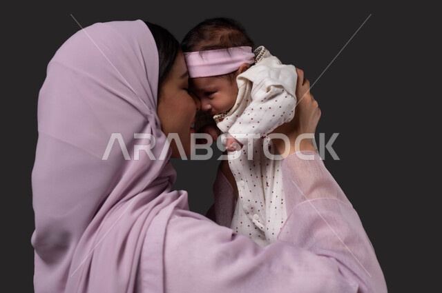 حنان الأمهات و تحمل المسؤولية والعناية بالأطفال، ملاعبة الأطفال بعطف واهتمام، قضاء الوقت باللعب مع الأبناء بإيماءات السعادة والسرور،  بورتريه مقرب من الجانب لأم عربية خليجية سعودية تبتسم ترتدي العباءة تحمل طفلتها الرضيعة وتقوم بمداعبتها بحب وحنان, خلفية سوداء